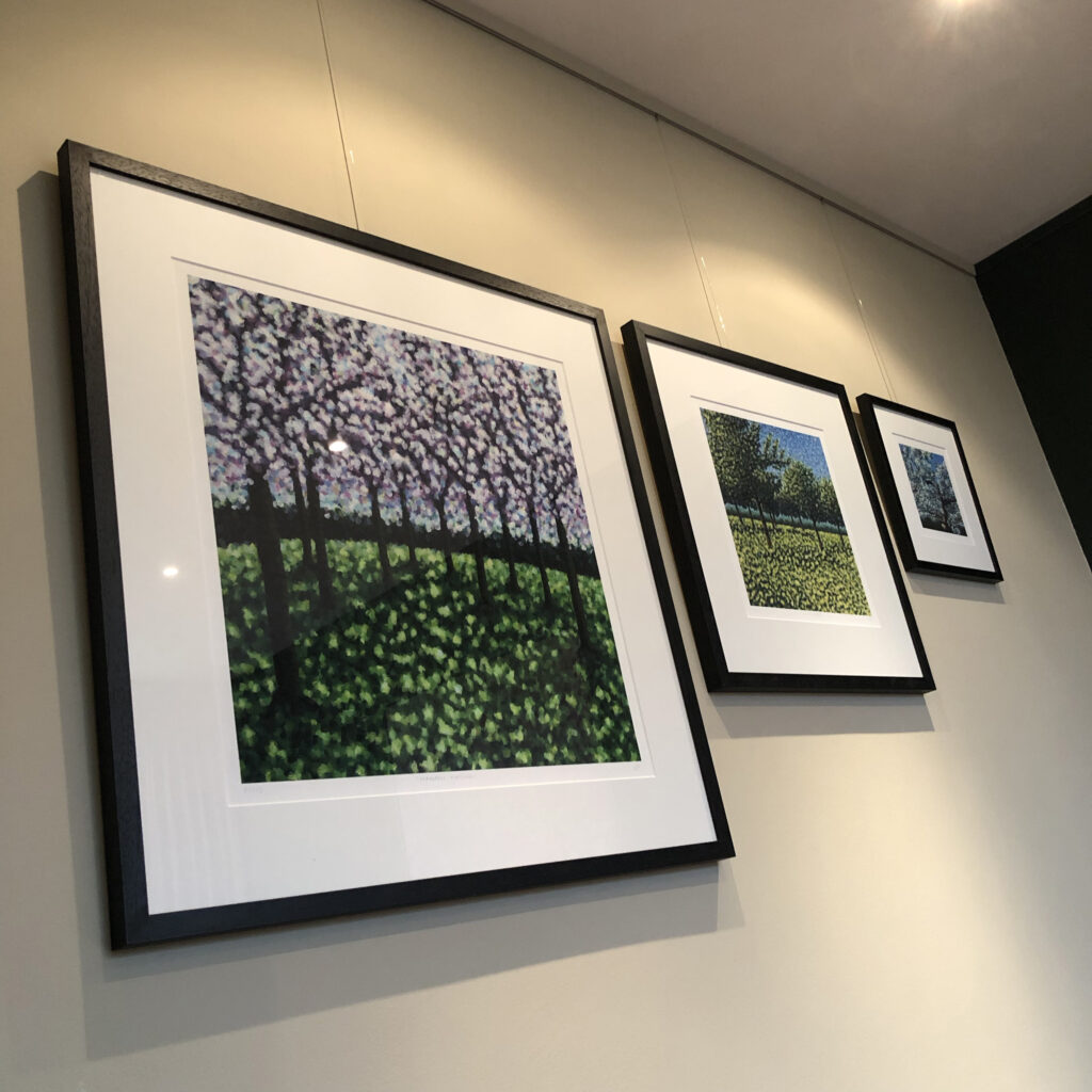 Drie ingelijste kunstwerken met kleurrijke landschapsschilderijen hangen aan een beige muur in een galerijopstelling.